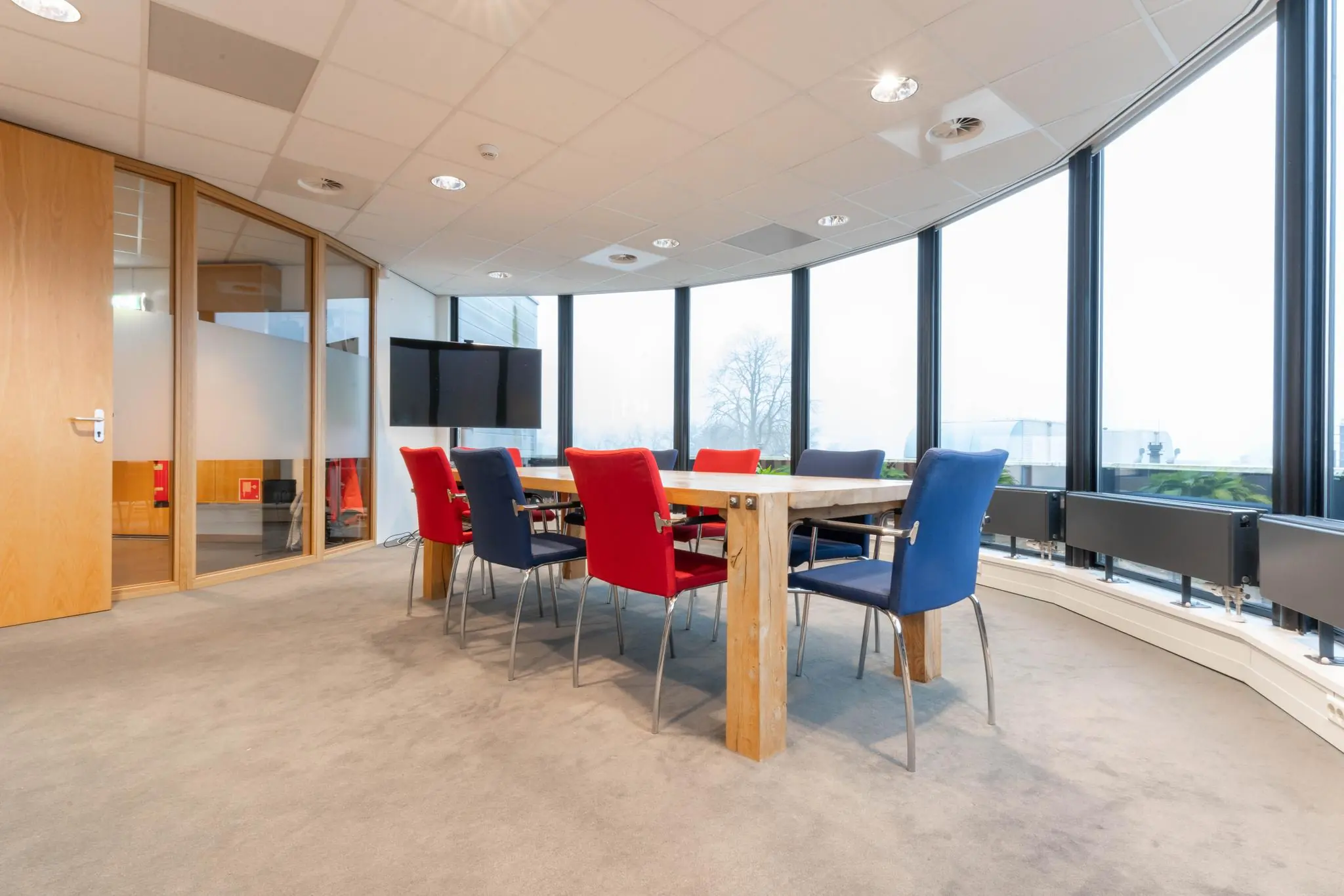 Vergaderruimte met houten tafel, rood-blauwe stoelen en grote ramen in het kantoor aan de 1e Hogeweg.